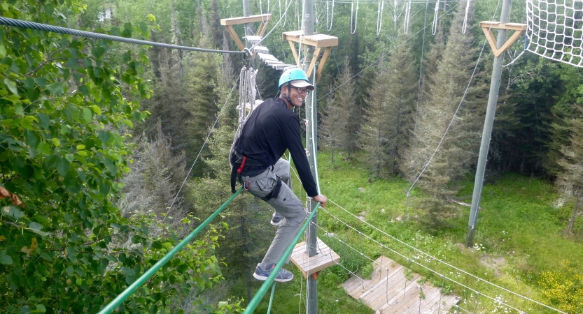 A person wearing safety gear and secured by ropes balances on an obstacle on a high ropes course. 